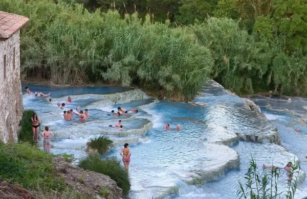 Persone si rilassano nelle cascate naturali dei aspio terme, con vegetazione lussureggiante e un antico edificio in pietra.