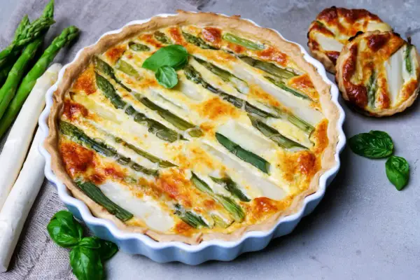 Herzhafte Spargel Quiche mit frischem Spargel und Kräutern, goldbraun gebacken. Daneben liegen roher Spargel und kleine Quiches.