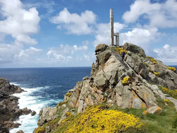 Escaleras de piedra ascienden por rocas cubiertas de flores amarillas hasta un monumento en el cabo Priorino. El mar azul y el cielo con nubes completan la escena.