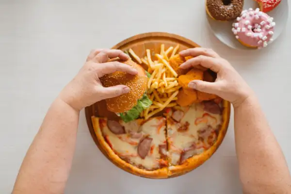 Mani che afferrano un hamburger, patatine, nuggets e pizza. Un vero **cheat day** con ciambelle in lontananza.