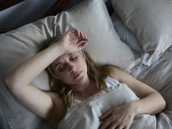 Mujer de unos 40 años sufre sudores nocturnos, con la mano en la frente, en la cama.