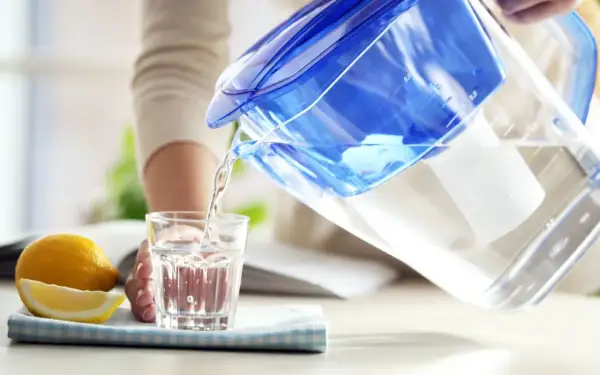Une personne verse de l'eau filtrée d'une carafe bleue dans un verre. À côté, un citron coupé. L'eau filtrée est elle bonne pour la santé ?