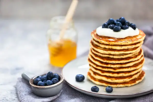 Pile de pancakes moelleux, garnis de crème et de myrtilles, avec un pot de miel en arrière-plan. Une recette pancake parfaite pour le petit-déjeuner.