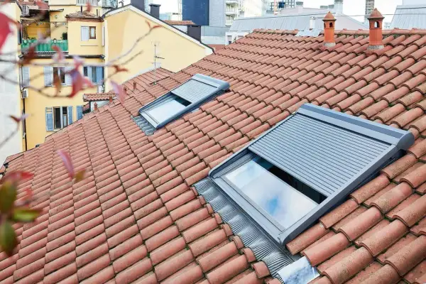 Tegole rosse su un tetto con una pendenza tetto 30 gradi, due lucernari con tapparelle grigie parzialmente abbassate.
