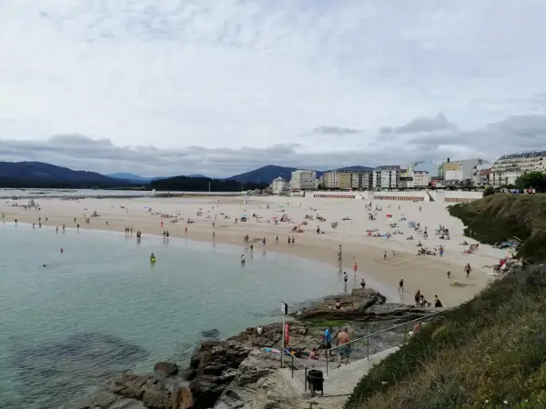 Gente disfruta de un día soleado en la playa de peizas, con aguas cristalinas y edificios al fondo.