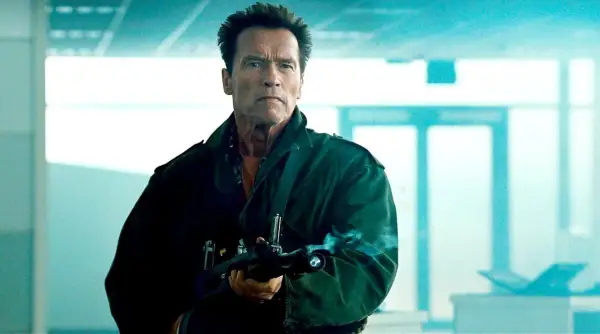 Arnold Schwarzenegger w zielonej kurtce z karabinem, gotowy do akcji w jednym ze swoich kultowych filmów.