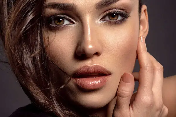 Primo piano di una donna con un sofisticato trucco occhi neri, labbra lucide e capelli castani mossi.