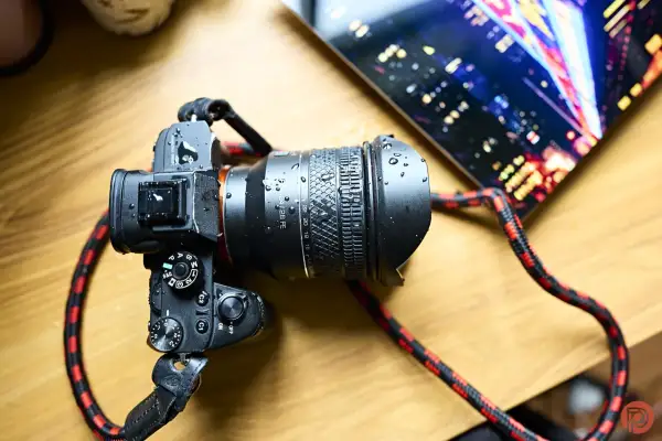A Rokinon 24-28mm FE lens on a camera, covered in water droplets. The camera has a red and black strap. A tablet with a city lights display is in the background.