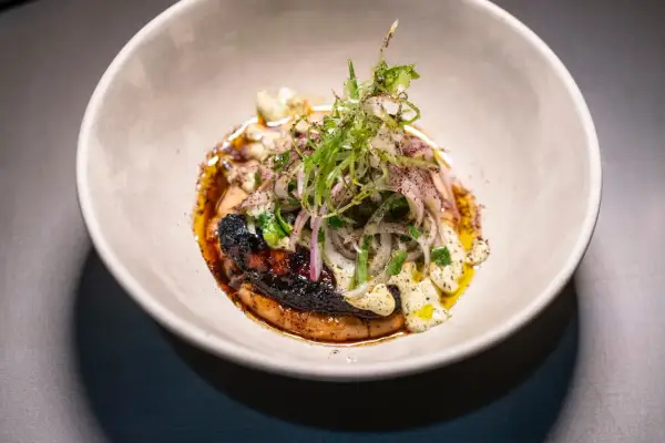 Plato gourmet con un trozo de carne oscuro y crujiente, cubierto de cebolla morada y hierbas frescas, bañado en una salsa dorada. Un solete Repsol para el paladar.
