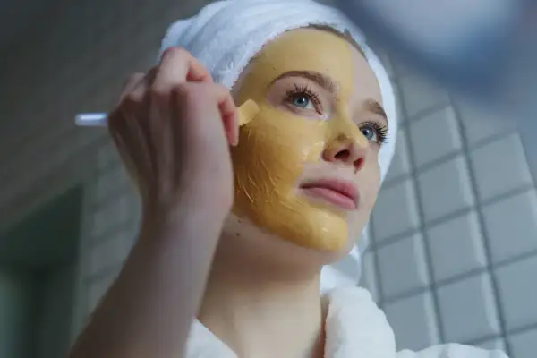 Junge Frau mit Handtuch auf dem Kopf trägt eine Kurkuma-Maske auf. Vorher-Nachher-Effekt für strahlende Haut.
