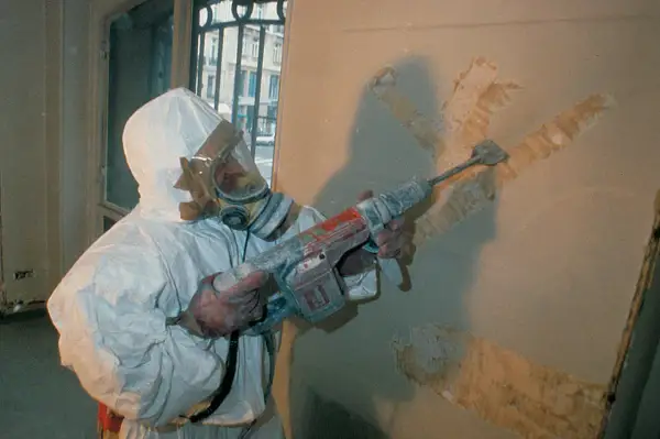 Travailleur en combinaison intégrale et masque à gaz, utilisant un marteau-piqueur pour retirer un revêtement mural. Que faire face à l'amiante au travail.