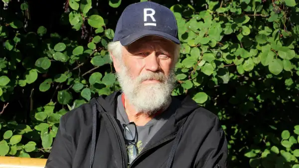 En äldre man med vitt skägg och keps med ett R på sitter utomhus. Han är en del av Ronny Eriksson familj.