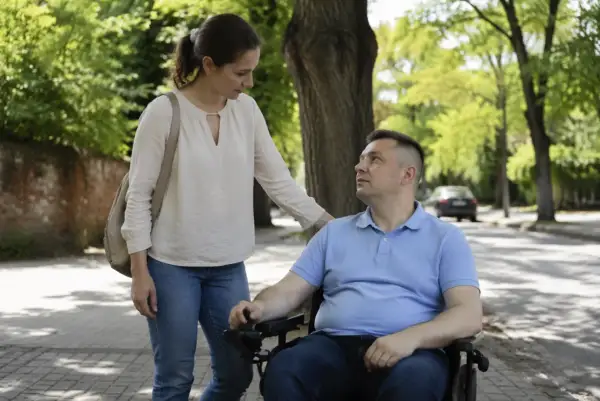 Kobieta wspiera mężczyznę na wózku inwalidzkim na tle zieleni.