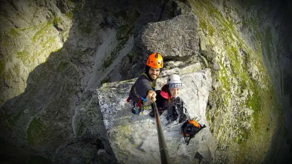 Uśmiechnięci alpiniści na skalnej półce, gotowi na wejście na mnicha. Widok z góry na stromą ścianę.