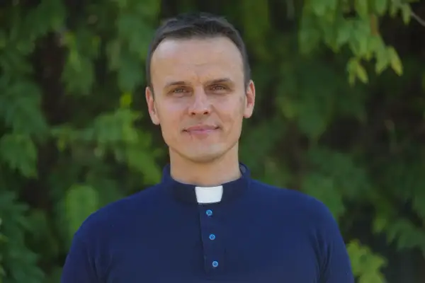 Młody ksiądz z osiedla w granatowej koszuli z kołnierzykiem, uśmiecha się na tle zieleni.