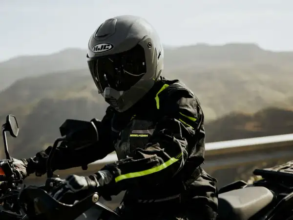Motocyklista w szarym kasku HJC i czarnej kurtce z neonowymi detalami jedzie motocyklem po górskiej drodze.
