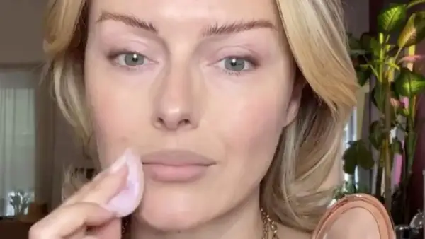 Eine Frau trägt mit einem Schwämmchen puder auf ihr Gesicht auf. Ihr blondes Haar umrahmt ihr Gesicht, während sie sich auf ihr Make-up konzentriert.