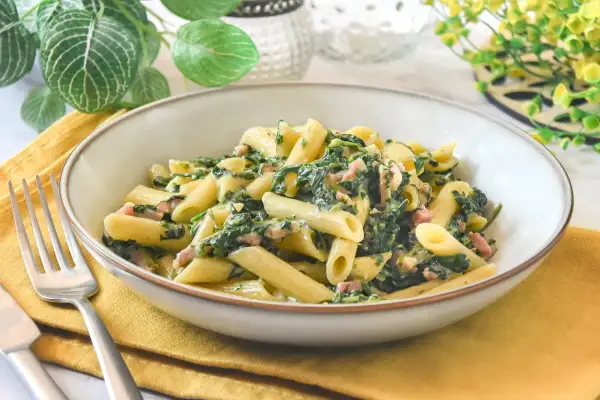 Un plato de pasta penne con espinacas y trocitos de jamón, servido en un cuenco sobre una servilleta amarilla.