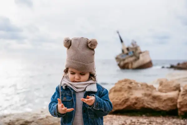 Małe dziecko w czapce z pomponami bawi się na plaży na Cyprze, w tle wrak statku.