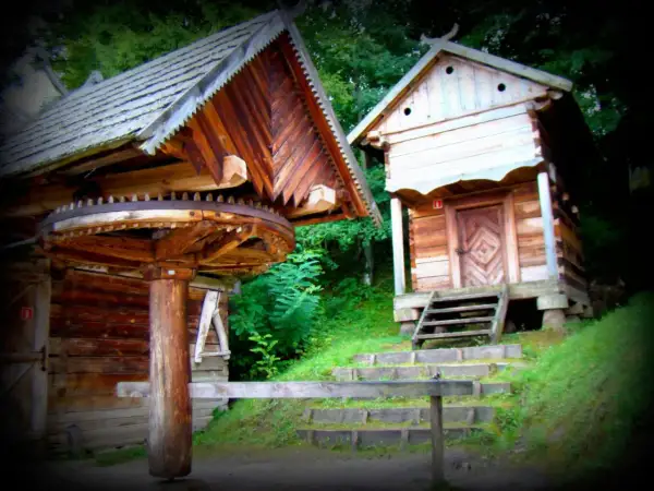 Drewniane chaty w skansenie, przypominające architekturę Kurpiów, z widocznym kołem młyńskim.