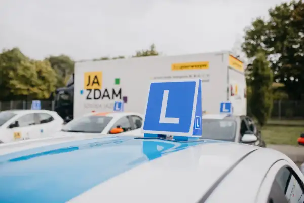 Na dachu białego samochodu nauki jazdy stoi niebieska tablica z literą "L". W tle ciężarówka z napisem "JA ZDAM". Ile można mieć błędów na egzaminie teoretycznym?
