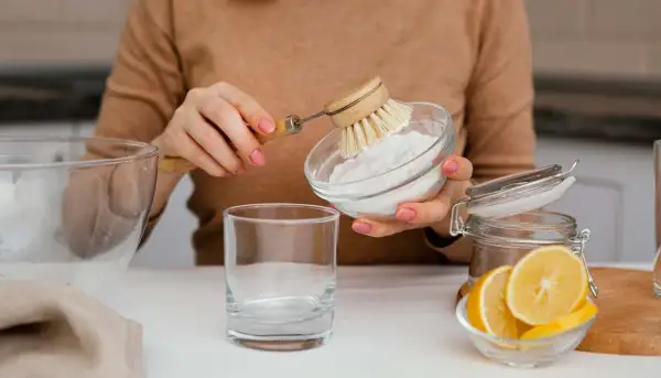 Manos con manicura rosa preparan una mezcla de agua con bicarbonato para limpiar. Se ve un vaso, limón y un cepillo.