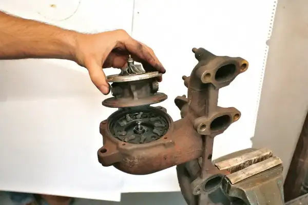 Dłoń trzyma turbosprężarkę, prezentując jej turbinę i wirnik. Część turbosprężarki jest zamontowana w imadle.