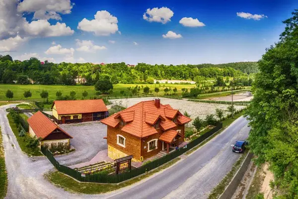 Drewniane budynki rancza z czerwonymi dachami, otoczone zielenią, pod błękitnym niebem z chmurami.