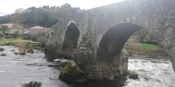 El histórico puente Maceira cruza un río caudaloso, con casas de tejados rojos y árboles frondosos al fondo.