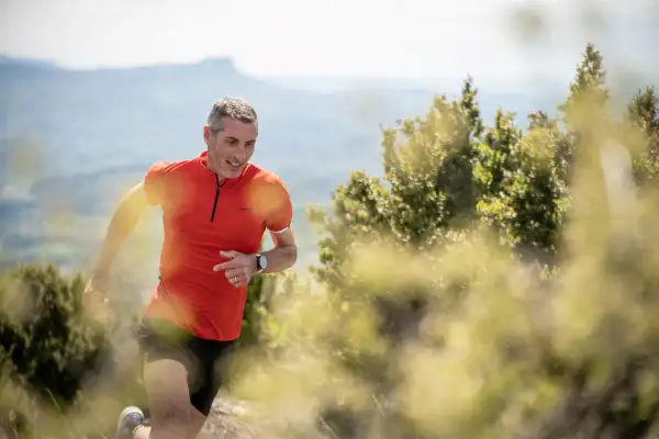Un homme en tenue de trail run, souriant, court sur un sentier de montagne.