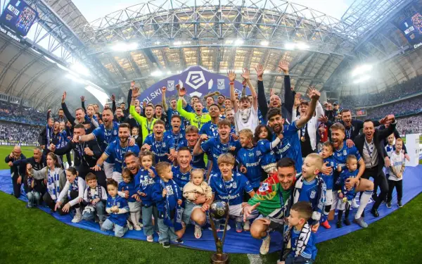 Byli piłkarze Lecha Poznań świętują z pucharem i dziećmi na murawie stadionu.