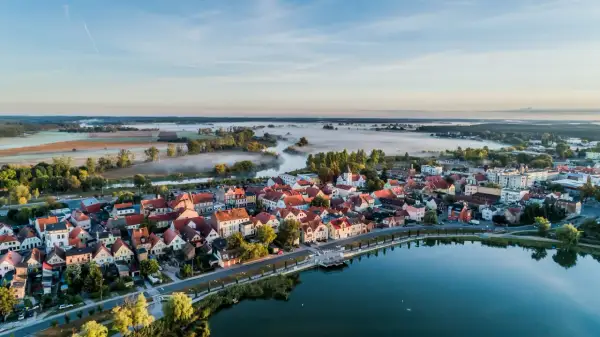 Poranna mgła nad rzeką i polami, otaczająca urokliwe miasteczko z czerwonymi dachami. To prawdziwa kraina 100 jezior, gdzie spokój miesza się z pięknem natury.