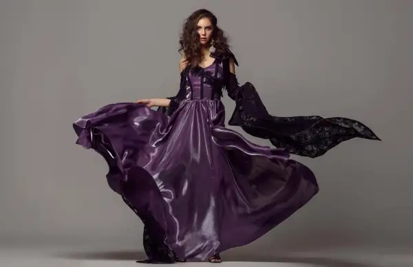 A model in a flowing purple gown and black lace sleeves poses for studio fashion photography.