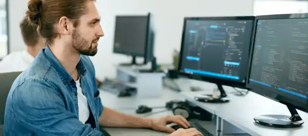 Manlig IT-tekniker med uppsatt hår och skägg arbetar vid två datorskärmar med kod.