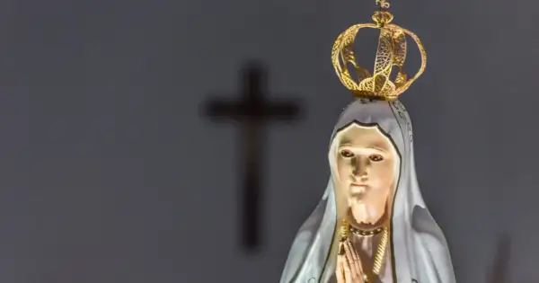 Figurka Matki Bożej z Fatimy, w koronie, z modlącymi się dłońmi. W tle niewyraźny krzyż, symbol objawienia fatimskiego.