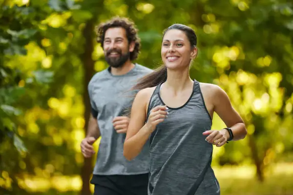 Coppia sorridente corre nel parco. Lei, in primo piano, è determinata, lui la segue con gioia. Un esempio di come iniziare a correre a 50 anni sia possibile e appagante.