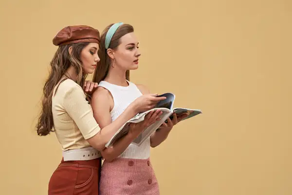 Dwie kobiety w stylizacjach inspirowanych **modą lat 60.** przeglądają magazyn. Jedna w skórzanym beretku, druga w opasce na włosach.