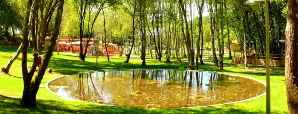 Un estanque circular refleja los árboles en un parque con un área de juegos y mesas al aire libre. Un lugar perfecto para un paseo tranquilo.