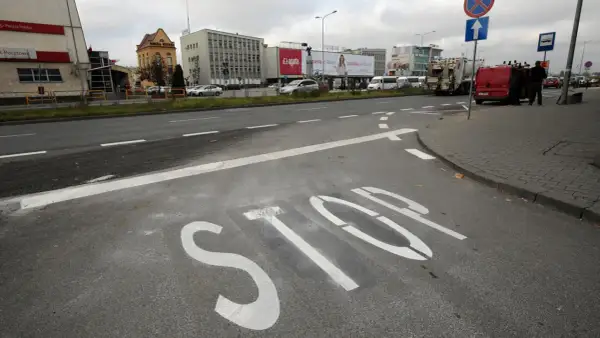 Na asfalcie widoczna jest biała linia warunkowego zatrzymania "STOP". W tle budynki i ruch uliczny.