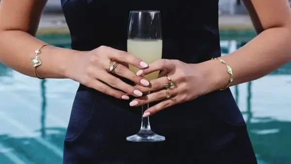 Manos con uñas rosadas y varios anillos dorados, sosteniendo una copa de champán. Brazaletes dorados adornan ambas muñecas.