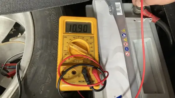 Multímetro amarillo muestra 10.90V al comprobar bateria coche. Se ve la batería, un neumático y cables.