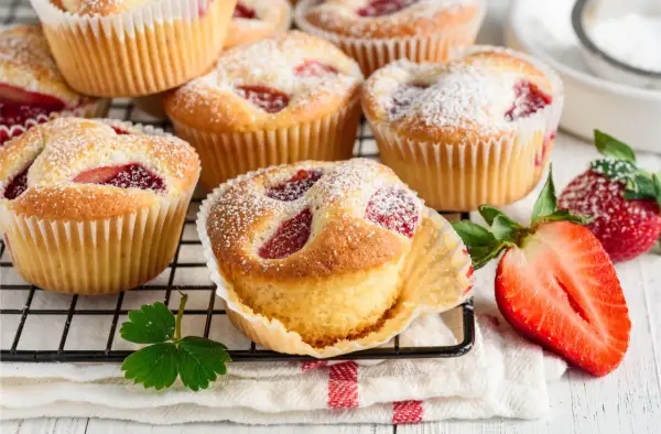 Puszyste muffiny z truskawkami, posypane cukrem pudrem, prezentują się apetycznie na kratce. Obok leżą świeże truskawki.