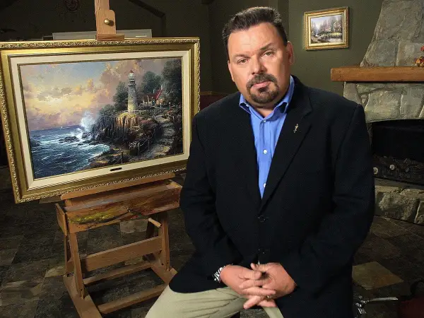 A man in a suit sits beside a Thomas Kinkade painting of a lighthouse on a cliff.
