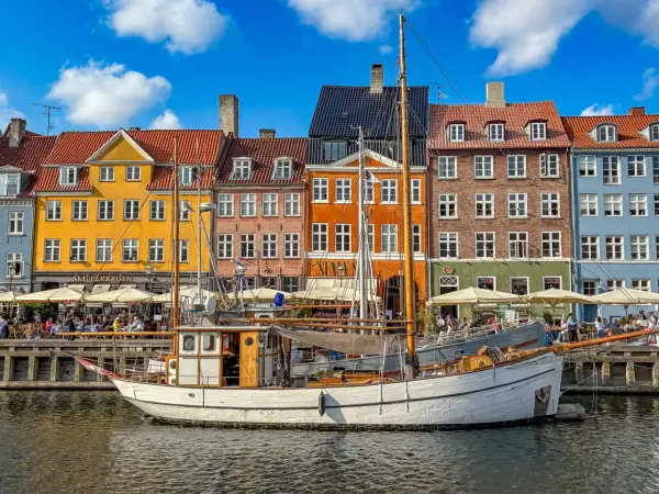 Ein weißes Segelschiff liegt im Hafen vor bunten Häusern. Menschen sitzen in Cafés am Wasser. Ein perfekter Tag im kopenhagen sommer.