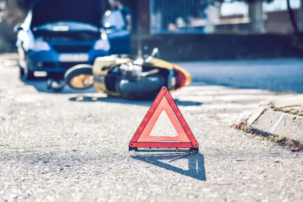 Trójkąt ostrzegawczy na drodze po wypadku. Złoty skuter leży na boku, obok niebieski samochód z otwartą maską. Może to być symbol problemów z ubezpieczeniem, jak 2 rata OC po terminie.