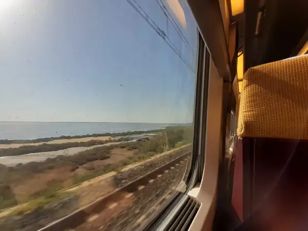 Blick aus dem Zugfenster auf eine Küstenlandschaft mit Meer und Bahngleisen. Die Reise mit dem Zug nach Mallorca beginnt.