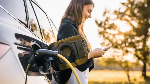 En kvinna laddar sin elbil utomhus, med solen som lyser genom träden. En gul laddningskabel är ansluten till bilen.