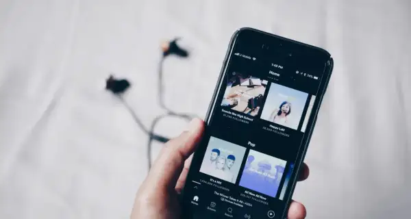Dłoń trzyma smartfon z aplikacją do streaming muzyki. W tle leżą słuchawki.