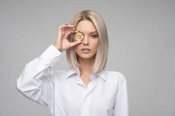 Blondynka w białej koszuli trzyma monetę bitcoin przed okiem, czując zazdrość, że ktoś ma lepiej.