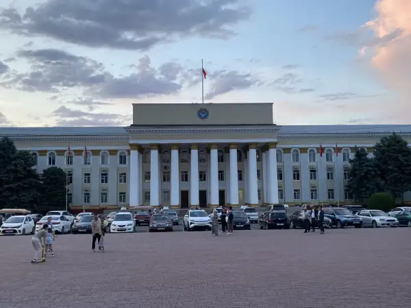 Plac przed budynkiem rządowym w Biszkeku, stolicy Kirgistanu. W tle monumentalna fasada z kolumnami, na dachu flaga. Na placu zaparkowane samochody i spacerujący ludzie.